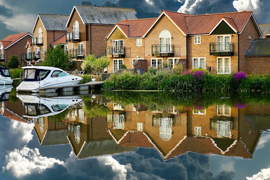A residential property situated along a canal features a brick-built house with large arched windows, some of which have small balconies with black metal railings. The house has a pitched roof with red tiles and is surrounded by greenery, including bushes and small trees. A wooden dock extends into the canal, on which a white motor boat with a covered cabin is moored, with its reflection visible in the calm water. The scene is set outdoors during daytime, with partly cloudy skies overhead. In the context of house removals, this image shows the type of home where movers like Movers Bow may assist clients with packing, furniture transport, and loading processes for a smooth relocation, whether inside or between properties near water or in similar residential areas.