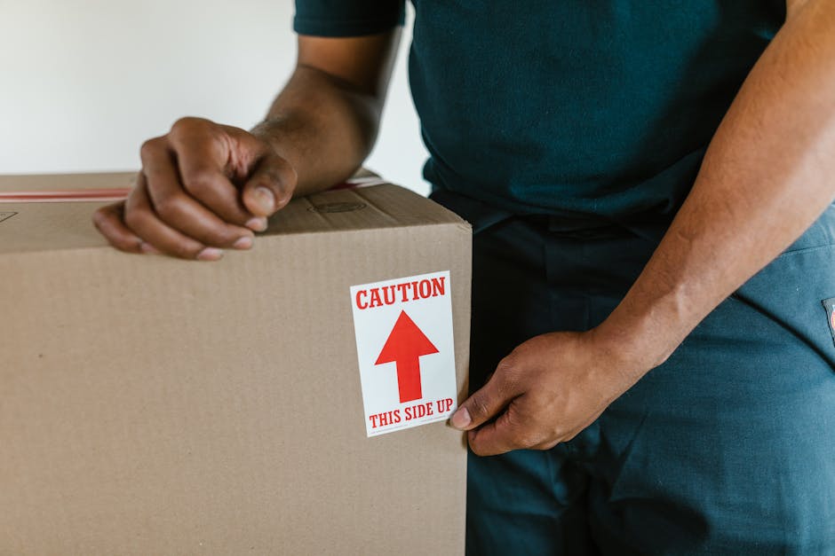 Close-up image of a person wearing a dark-colored shirt and blue trousers, handling a large cardboard box during a house removal. The individual is holding the box steady with one hand while the other hand is gripping the side of the box, which features a prominent red and white caution label with an upward arrow and the text 'CAUTION THIS SIDE UP'. The scene appears to take place indoors, with the individual positioned near a doorway or entrance, possibly preparing to load the box onto a moving van or carry it through a property. The cardboard box is sealed and wrapped with packing materials, indicating careful packing and handling as part of a furniture transport or packing and moving process. Equipment such as trolleys or blankets are not visible in this image, but the action suggests an organized and careful loading process, typical of professional removals services like those provided by Movers Bow as part of a home relocation or internal packing task.