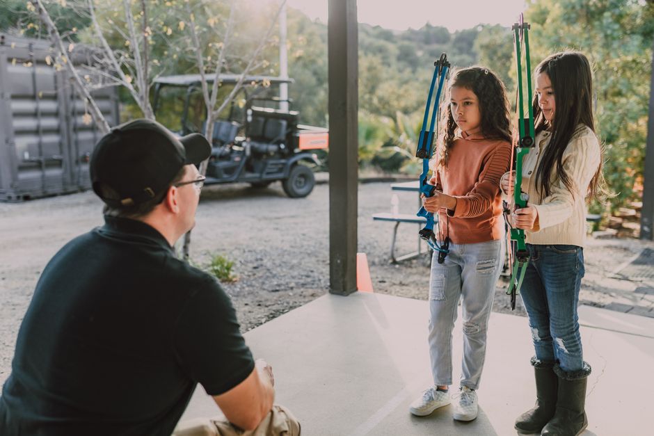A man wearing a black cap, glasses, and a black shirt is sitting on a concrete surface, observing two young girls holding colorful archery bows indoors near a large open door. The girls, one in a brown sweater and jeans, the other in a cream top and jeans with black boots, stand side by side facing the man, with the girl on the right holding a green bow and the girl on the left holding a blue bow, both appearing attentive. Outside, visible through the open door, there is an outdoor area with trees, a picnic table, and a parked utility vehicle, suggesting a rural or park setting. The scene shows natural daylight illuminating the space, which is part of a relocation or activity area, with elements like the concrete floor and the outdoor environment indicating a casual, recreational space suitable for workshops or team-building activities related to house removals or community moves. The scene’s focus on family-friendly activity supports the theme of move-related community or educational events coordinated by Movers Bow.