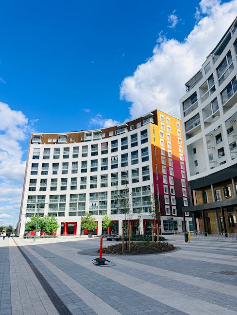 Photograph of a modern residential building situated in Queen Elizabeth Olympic Park, Bow, showing curved glass and concrete facades with multiple balconies. In the foreground, an outdoor loading area
